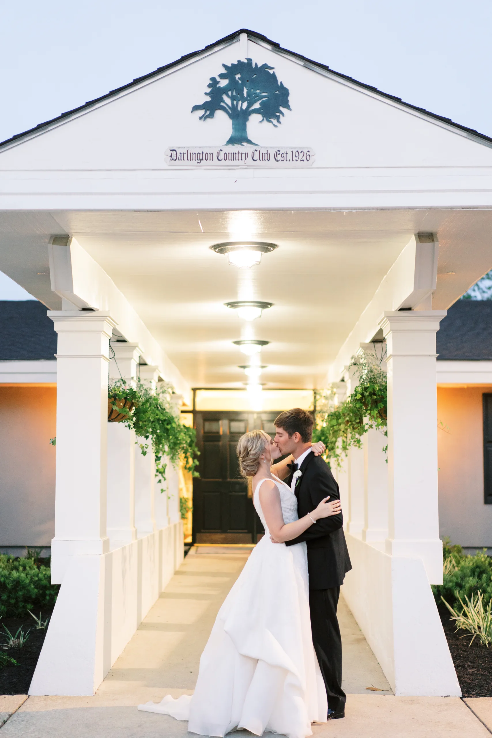 Bride and groom share a kiss outside the entrance to Darlington Country Club, lined with white columns.